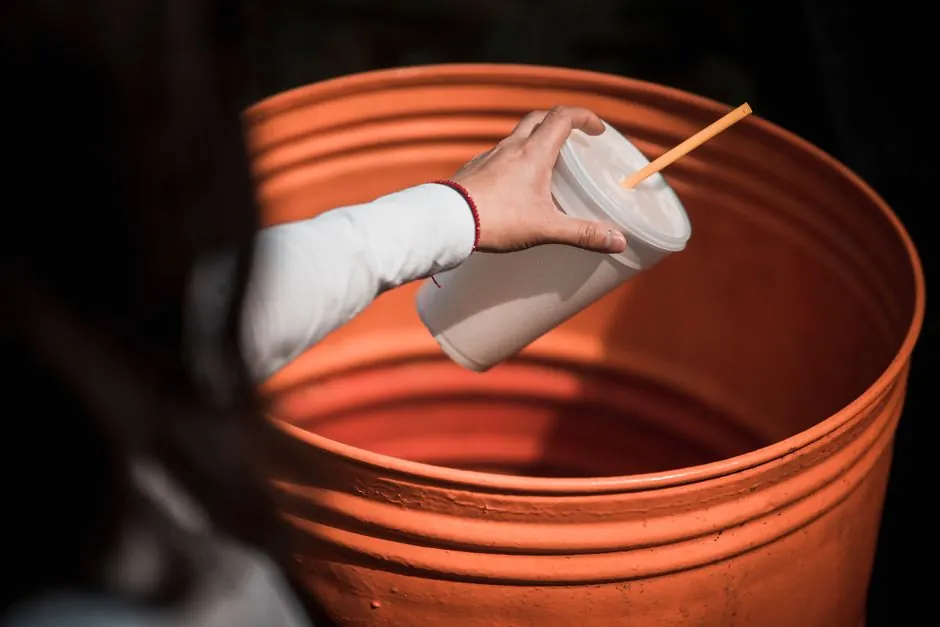 Close-up of a hand throwing a plastic cup into an orange trash bin, promoting recycling.